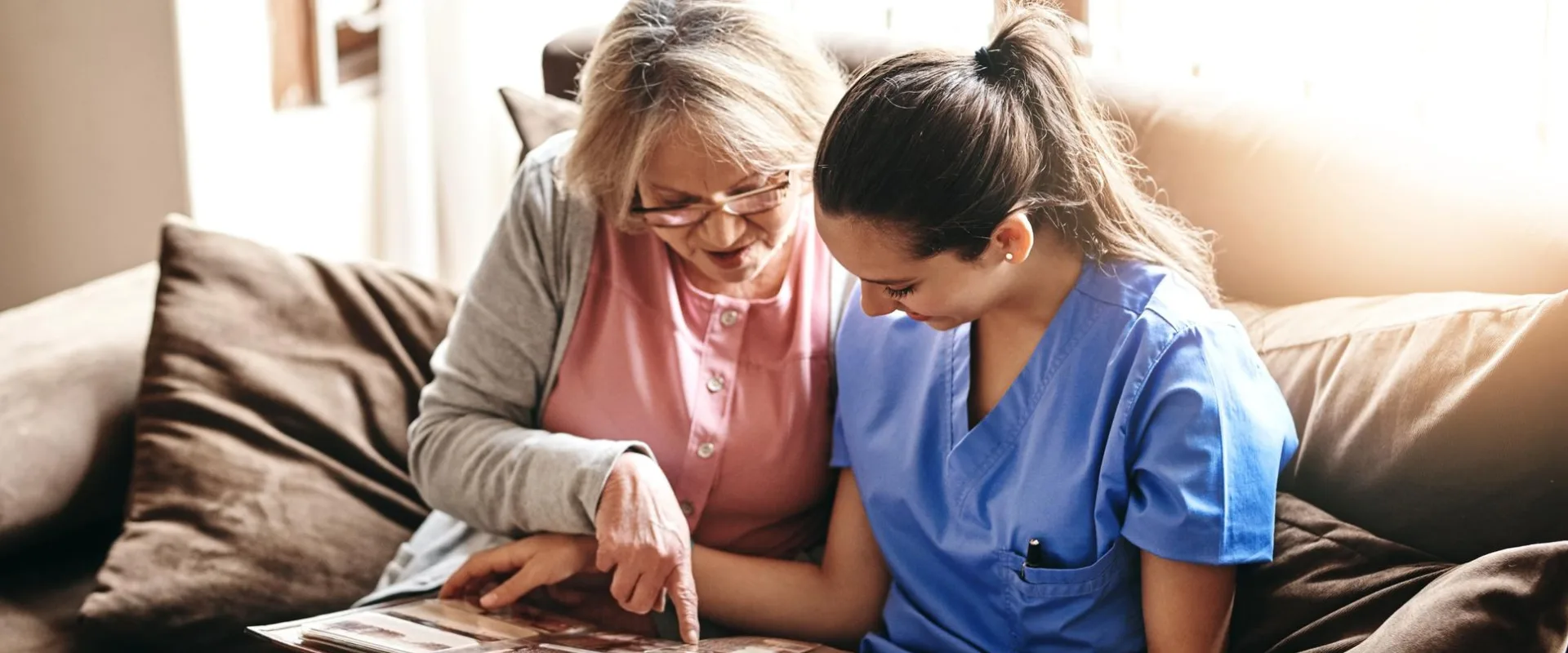 A woman provides companion care while seated with a patient on the couch through Thrive On Care.
